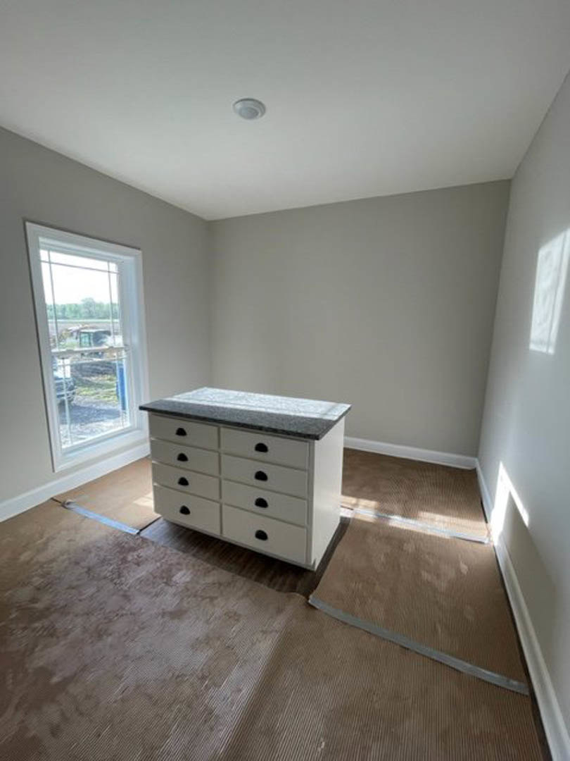 White cabinetry with black handles, grey laminate flooring, brown rug, large window with sheer white curtain, sunlight filtering into the room.