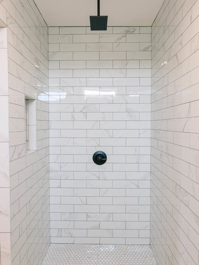 Shower with white hexagonal tile floor, white tile walls, black door handle, black square showerhead, and black rectangular drain cover