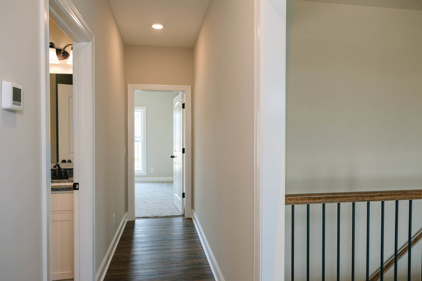Hallway with dark wood flooring, white baseboards, and a modern metal railing; open white door leads to a bright room with smooth plaster walls.