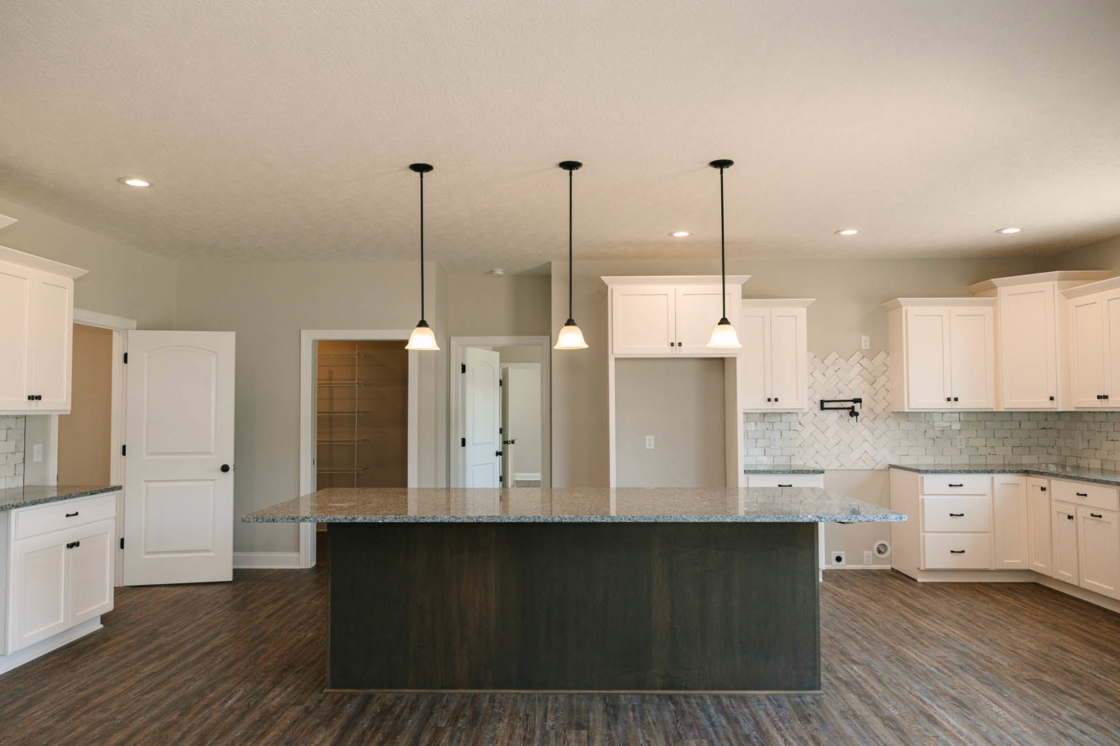 Spacious kitchen featuring a large central island with stone countertop, white cabinetry, tile backsplash, stainless steel sink, and black hardware accents
