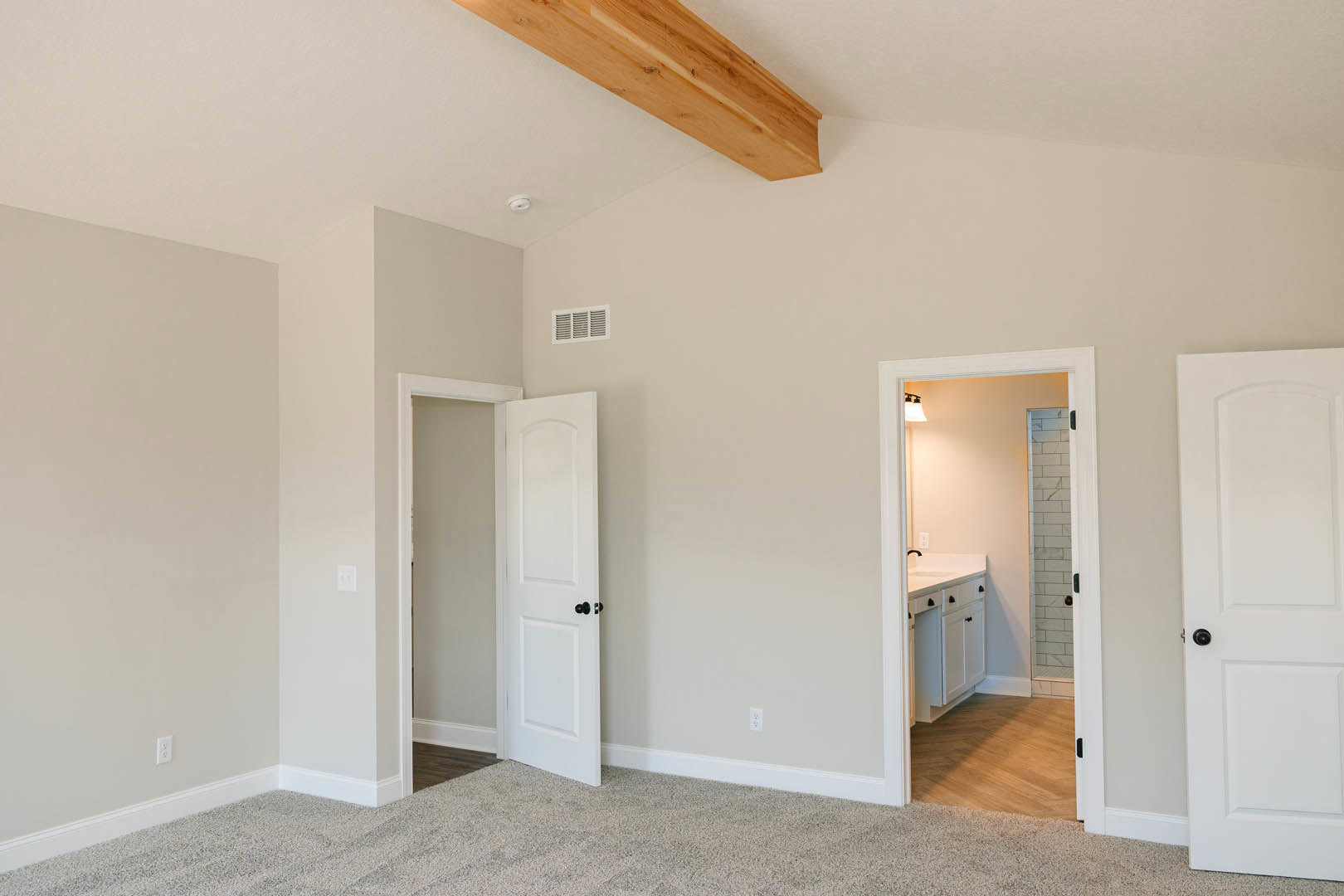 White paneled door with black hardware opening to a bathroom, wood beam ceiling, vent on light-colored wall, close-up view of natural wood flooring