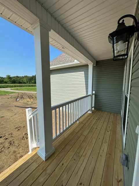 Wooden porch deck with white railing, grey house siding, and window; dirt yard in foreground.