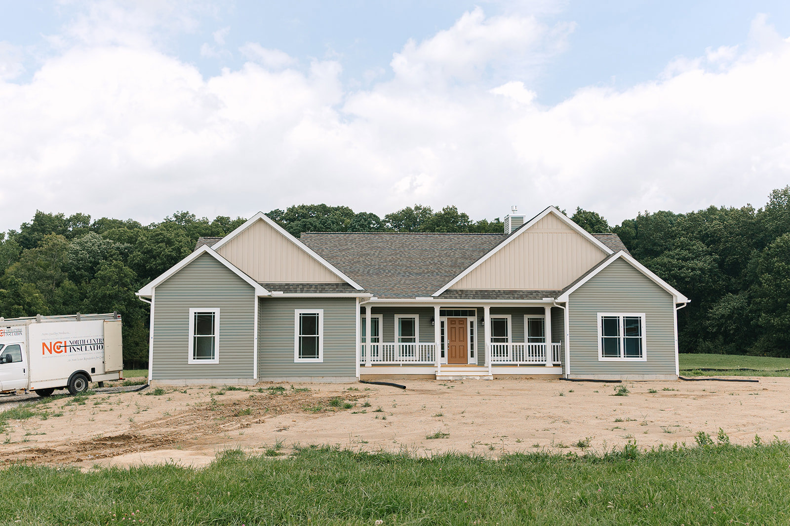 Single-story house with brown door and white trim, covered porch, white-framed windows, dirt yard, white truck parked in front, group of trees and cloudy sky in background