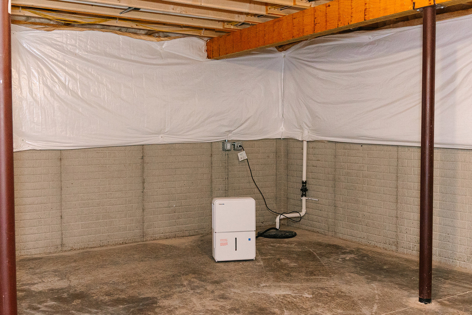 White rectangular utility box with small window mounted on basement wall, brown concrete floor, exposed ceiling with white sheet, visible white pipe and black wire, red support