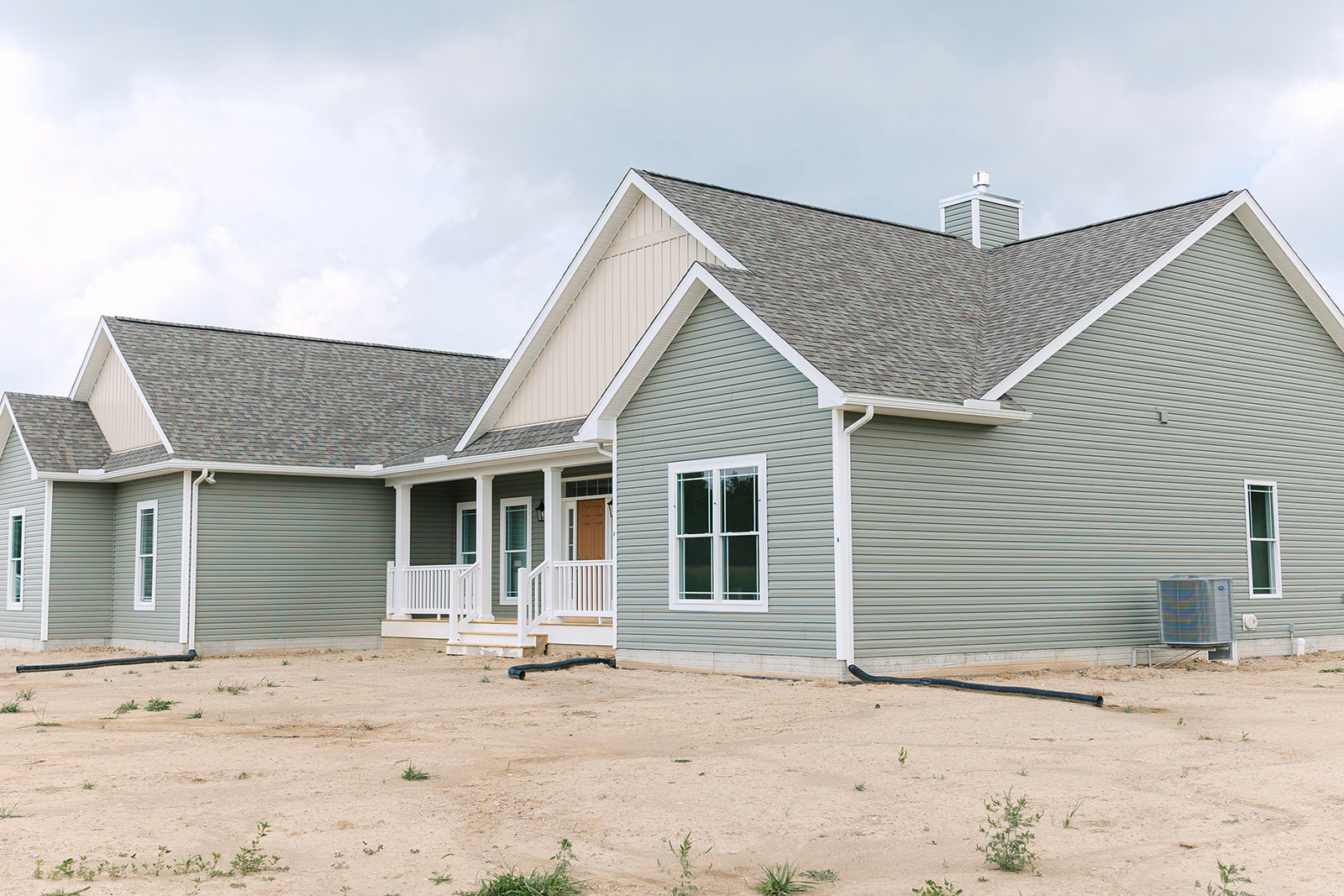 White-sided house with white railing, close-up window with white frame, dirt area in foreground featuring a black pipe and large metal utility box with cover, cloudy sky above.