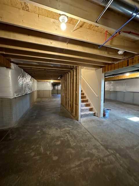 Concrete basement floor with unfinished stairs, exposed ceiling beams, white utility box, visible pipes, and plastered walls
