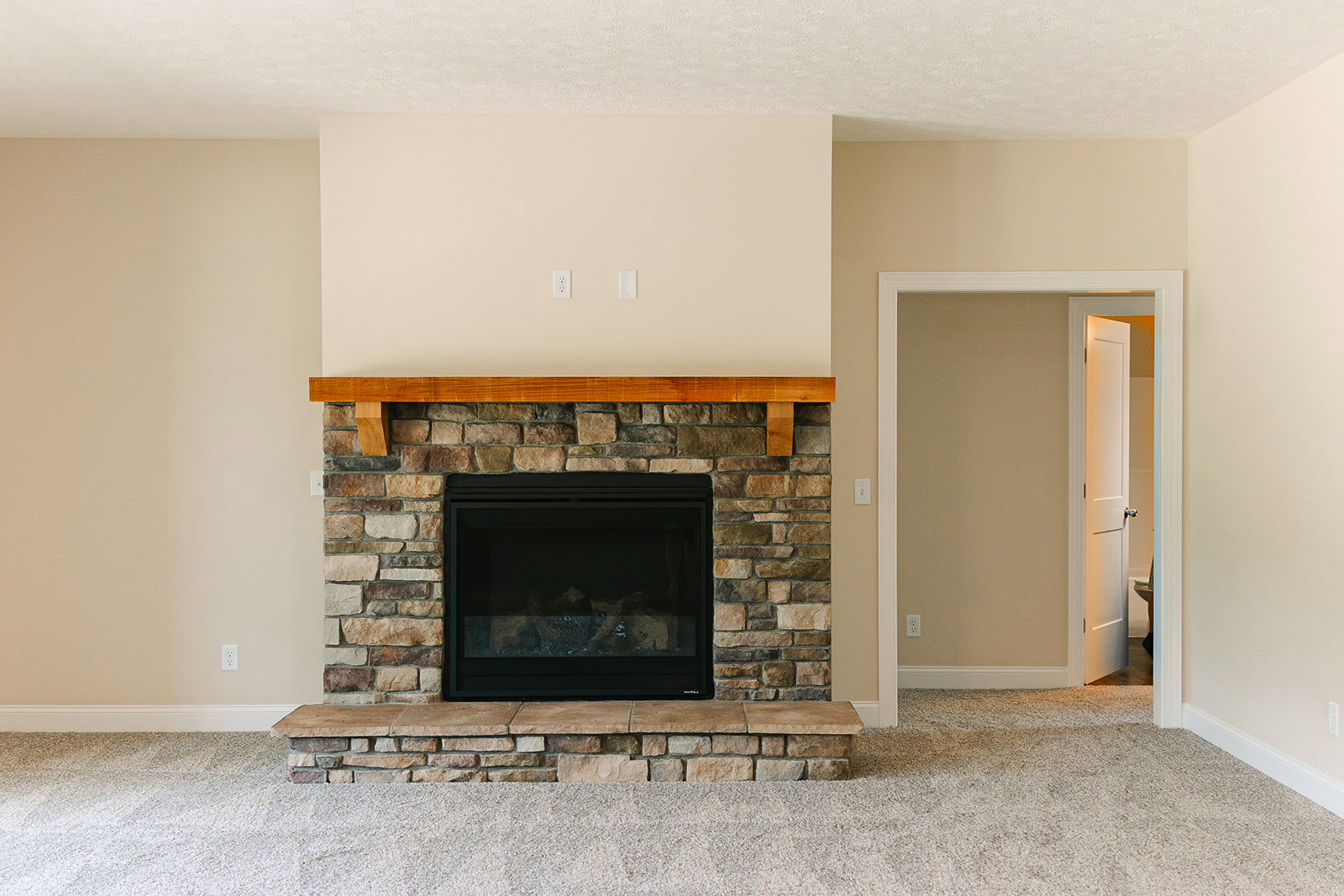 Stone fireplace with wood-burning stove, fire screen, and stone hearth in a living room; open door leads to adjacent space, white wall and black-and-white decor visible.