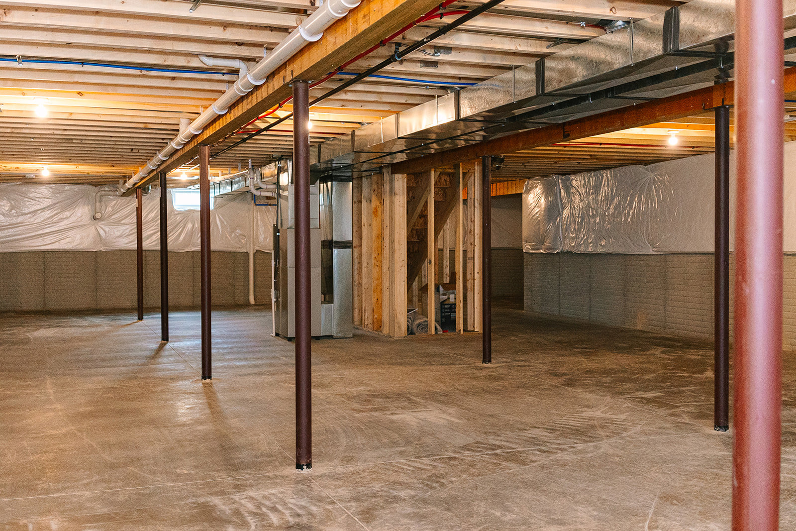 Exposed pipes along ceiling and walls, concrete floor, metal support pole, wooden shelf with rolled towels, brown flooring with black metal legs in unfinished basement room