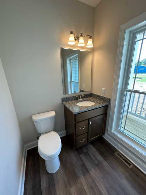 Modern bathroom with wood flooring, white toilet, stacked toilet seats, rectangular sink with cabinet, large wall mirror, and three-light fixture above.