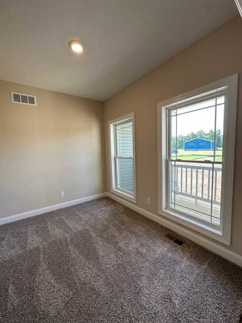 Carpeted room with white walls, large window with blinds, glass door opening to balcony and deck, blue shed visible outside, ceiling vent centered on wall