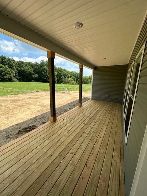 Wooden porch with green siding, white pillars, and deck overlooking grassy field and trees, window visible on house exterior
