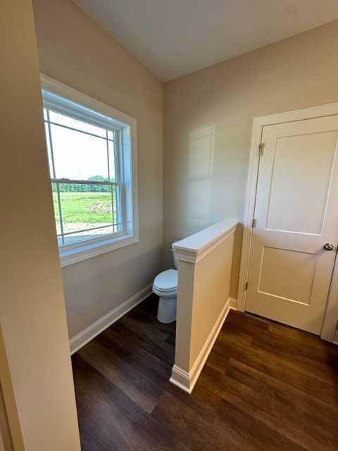 White toilet beside a window overlooking a grassy field, wood flooring with white trim, white wall, and a white door with a silver handle