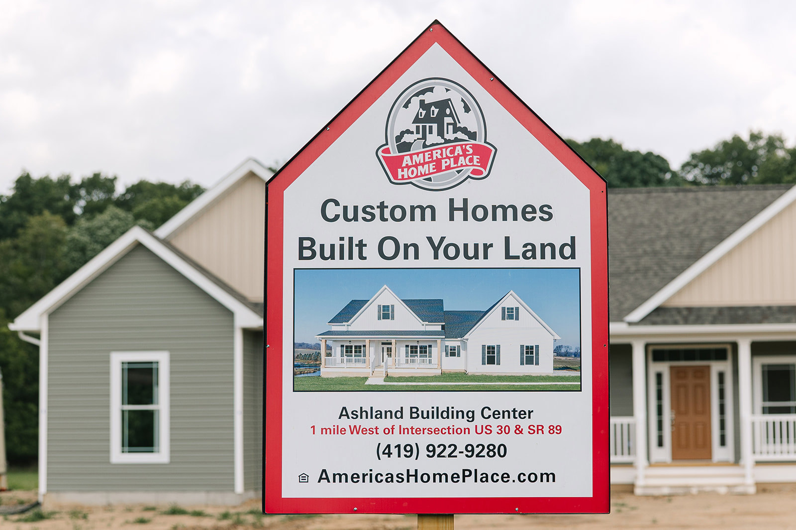 White real estate sign with house graphic stands on green lawn in front of two-story home with large windows, gray siding, and white trim; trees and cloudy sky in background
