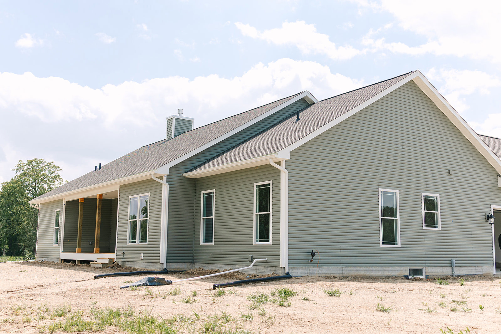 Partially built house with gray siding, white-framed windows, white gutters, shingled roof, brick chimney, and exposed dirt yard with a long black pipe lying on the ground.
