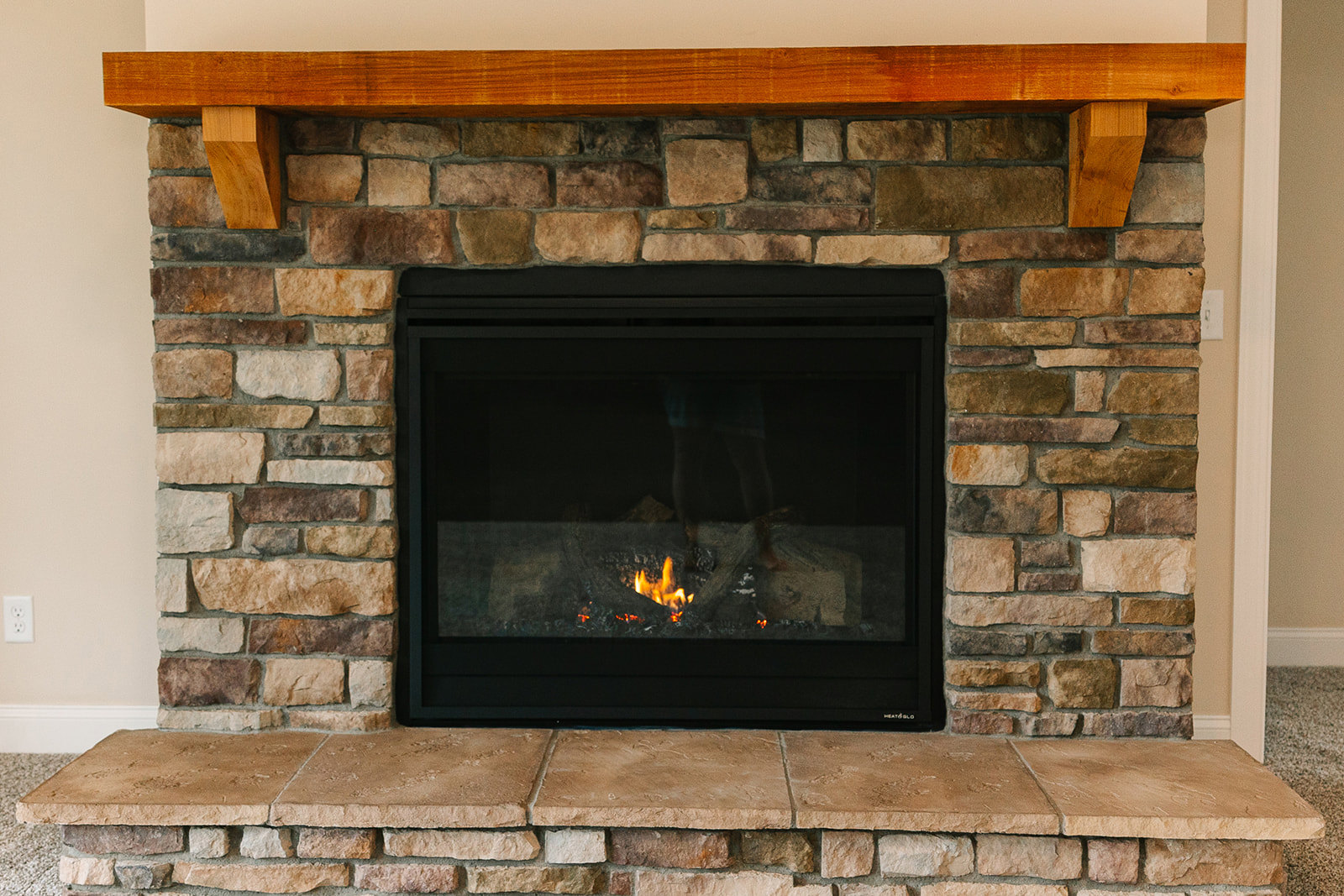 Stone fireplace with wood mantel, fire burning behind glass screen, brick hearth, and masonry wall in indoor living space
