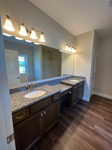 Bathroom with expansive mirror above double sinks, row of wall-mounted lights, wood flooring, white cabinetry, and chrome faucets
