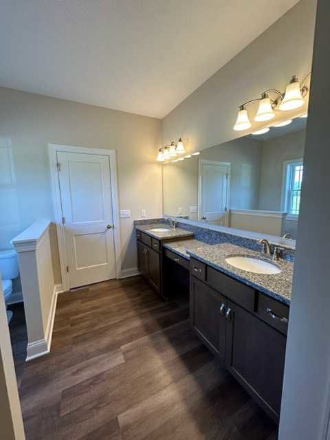 Bathroom with double sink vanity, white countertop, dark wood floor, white door with silver handle, light walls, recessed ceiling lighting