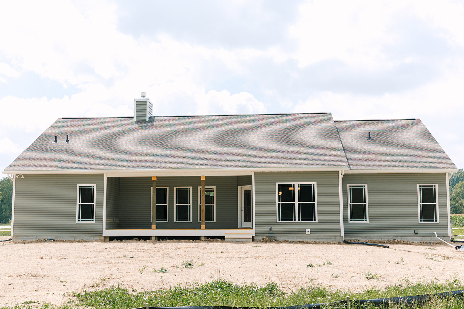 Modern home with white and black siding, large windows with white frames, covered porch, and unfinished dirt yard under a partly cloudy sky