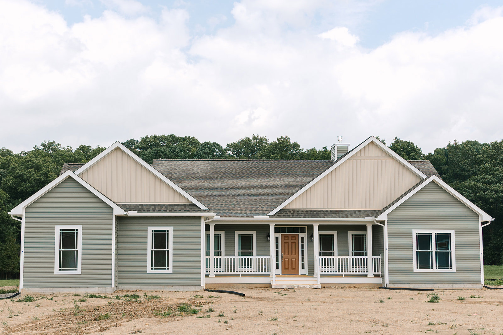 Two-story custom home with white-framed windows, covered front porch, and brown dirt yard bordered by tall trees under a partly cloudy blue sky