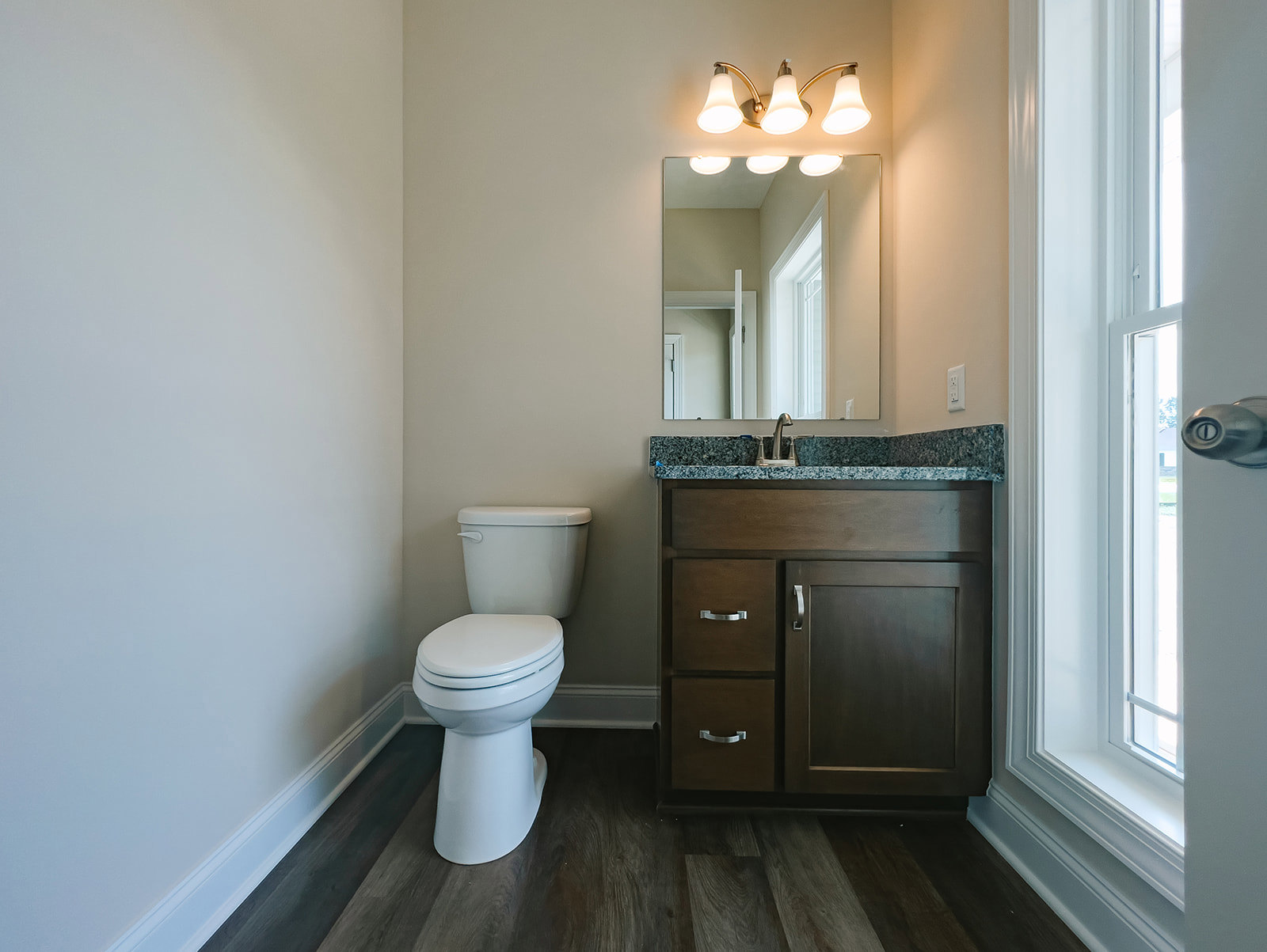 Bathroom with white toilet, black cabinet with handle, marbled countertop, sink, wall-mounted mirror with lights, and light fixture above.