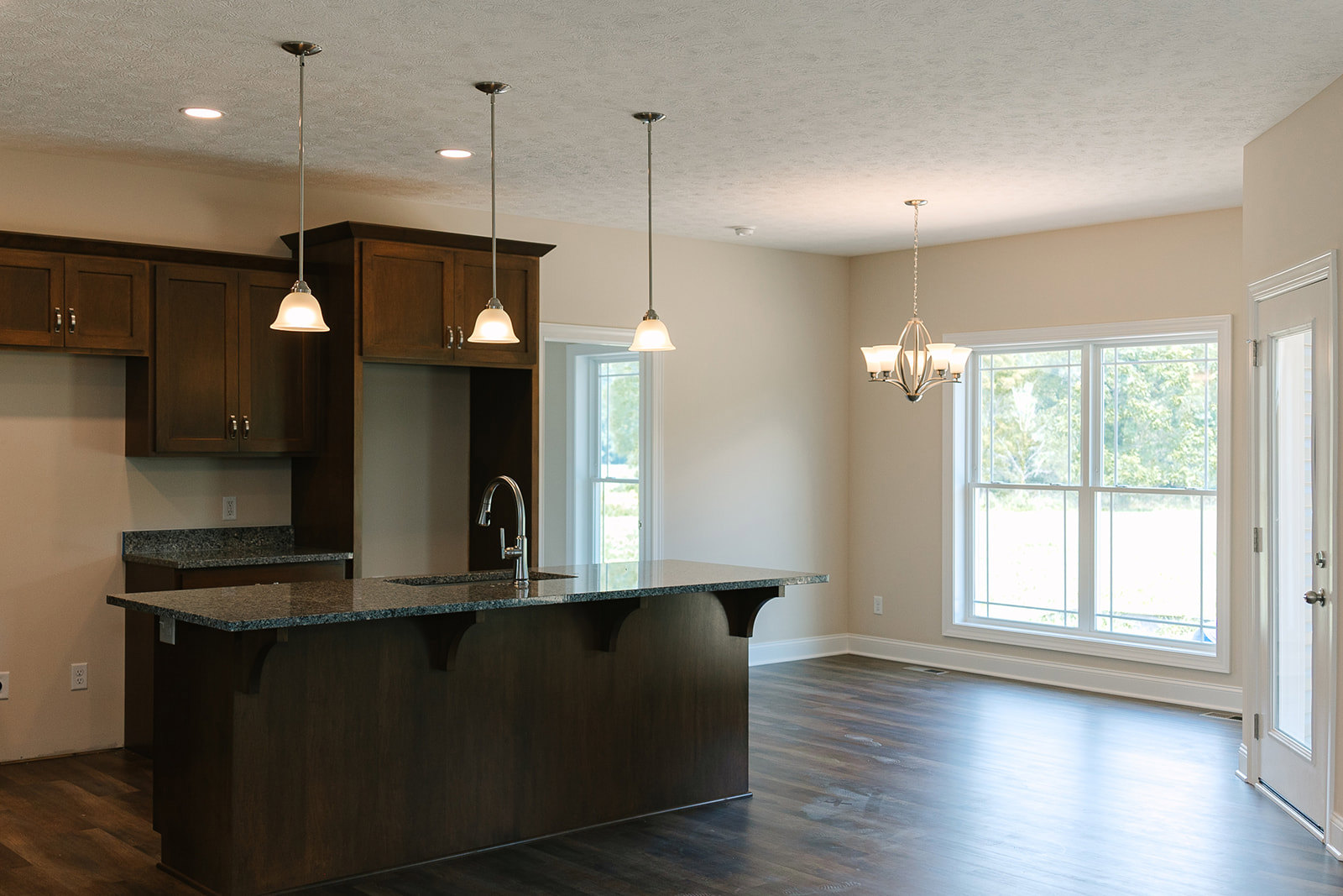 Modern kitchen featuring a central island with a built-in sink, white cabinetry, stone countertops, large window overlooking trees, wood flooring, and a ceiling-mounted chandelier.