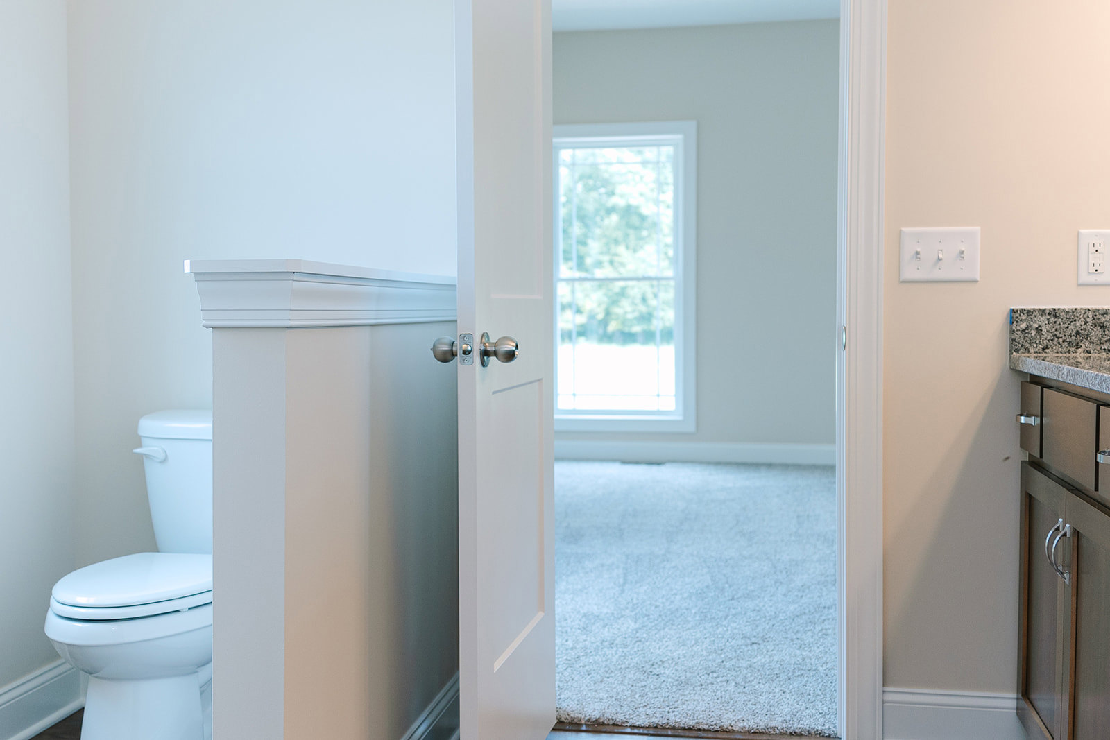 White door with metal handle open to a bathroom featuring white tile floor, close-up of toilet, white-framed window, light switch, and textured carpet adjacent to a white wall.