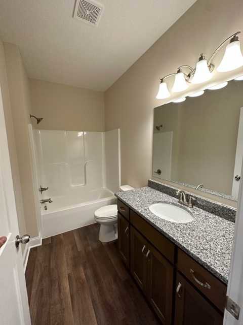 Bathroom with marble countertop, undermount sink, framed mirror, white trim, dark wood flooring, ceiling vent, multi-bulb light fixture, and visible toilet.