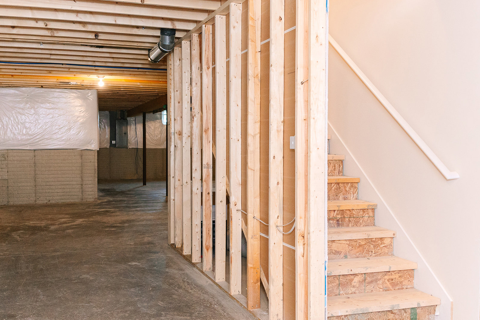 Wood-framed staircase under construction with exposed beams, chipboard side panels, plastic sheeting, and visible metal pipe in unfinished interior space