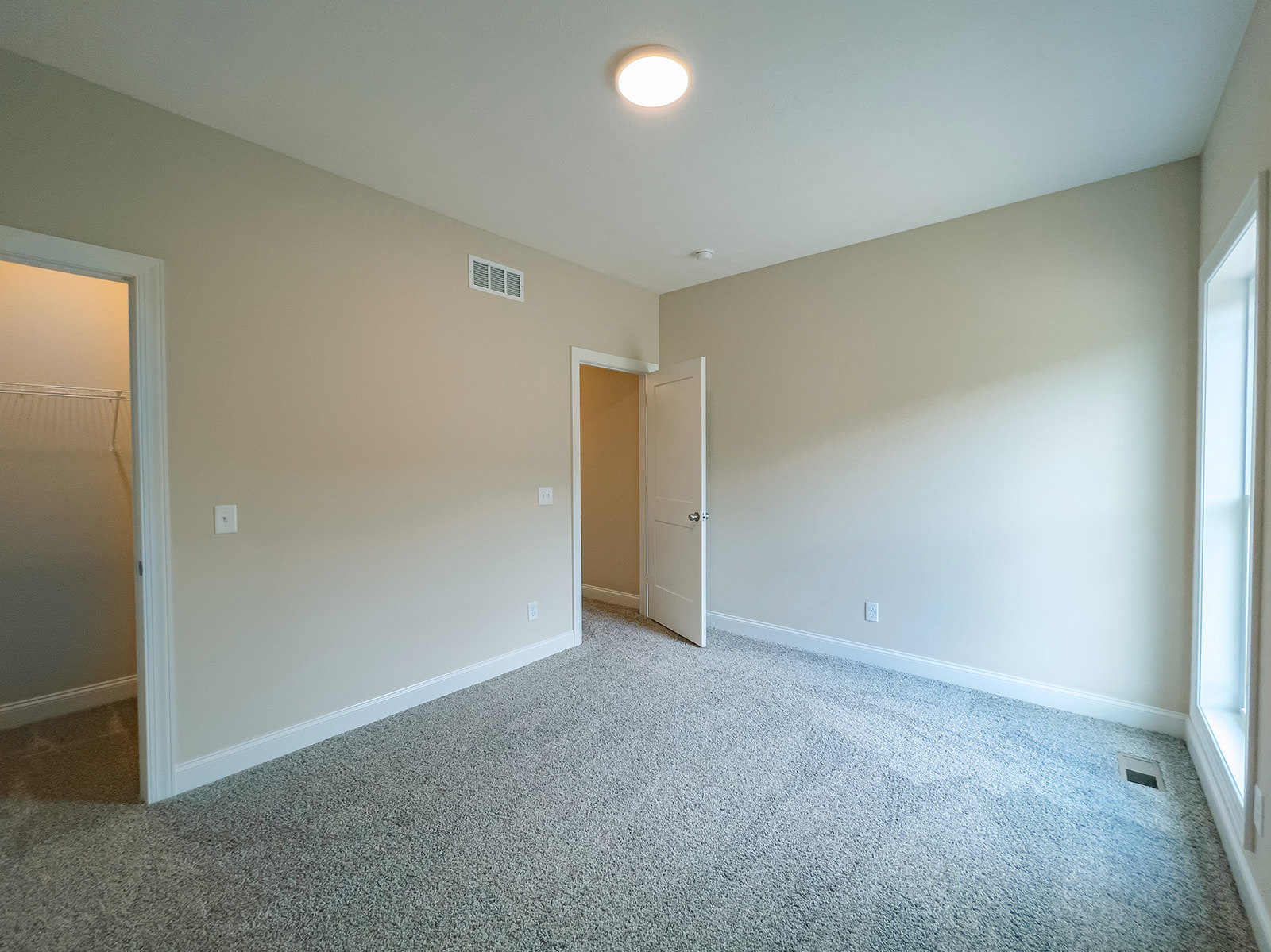 Carpeted room with white door open, silver knob, ceiling light fixture, wall vent, and window letting in natural light