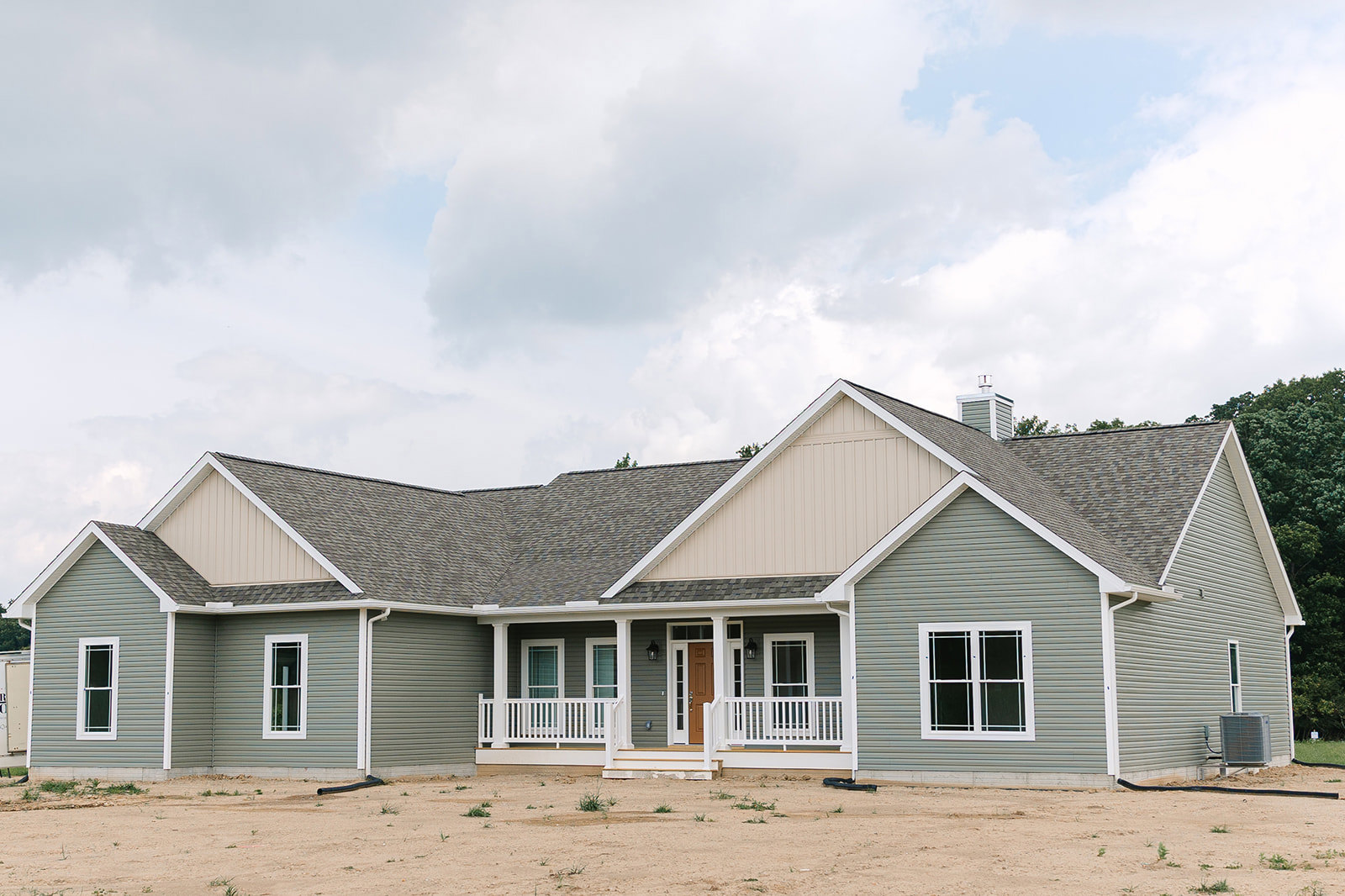 Single-story home with gray siding, white porch railing, black-framed windows, dirt yard, and rooftop HVAC unit with exposed wiring.