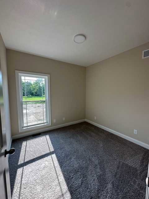 Bright room with white plaster walls, wood laminate flooring, carpeted area, white ceiling with recessed light, single door, large window overlooking grassy yard and trees
