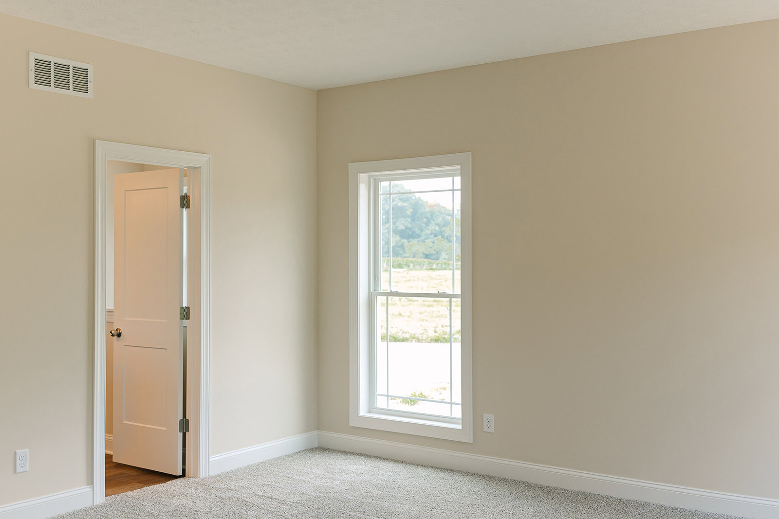 Carpeted room with white walls, large window overlooking grassy field, white door, wall vent, and baseboard molding