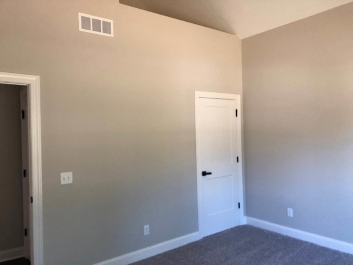 White paneled door with black handle set in a plaster wall, two white light switches, beige carpet flooring, and a rectangular air vent visible near the baseboard.