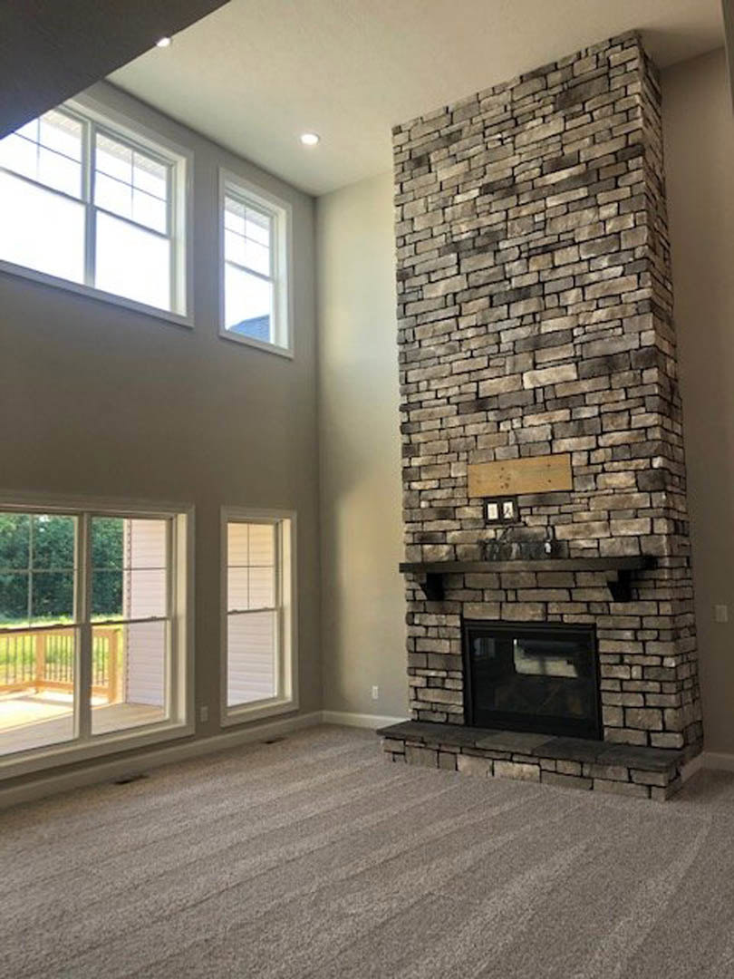 Stone fireplace with wood mantel and decorative sign, hardwood flooring, window with blinds, railing and deck visible through glass