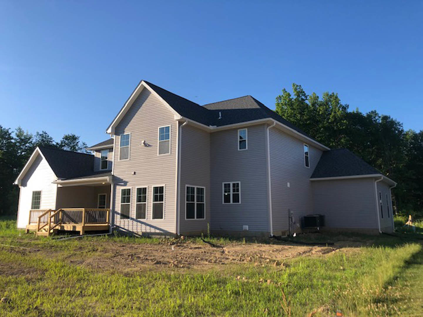 Two-story home with wood deck and railing, ramp leading to porch, patchy grass yard, multi-pane windows, blue sky overhead, small black object on outdoor table