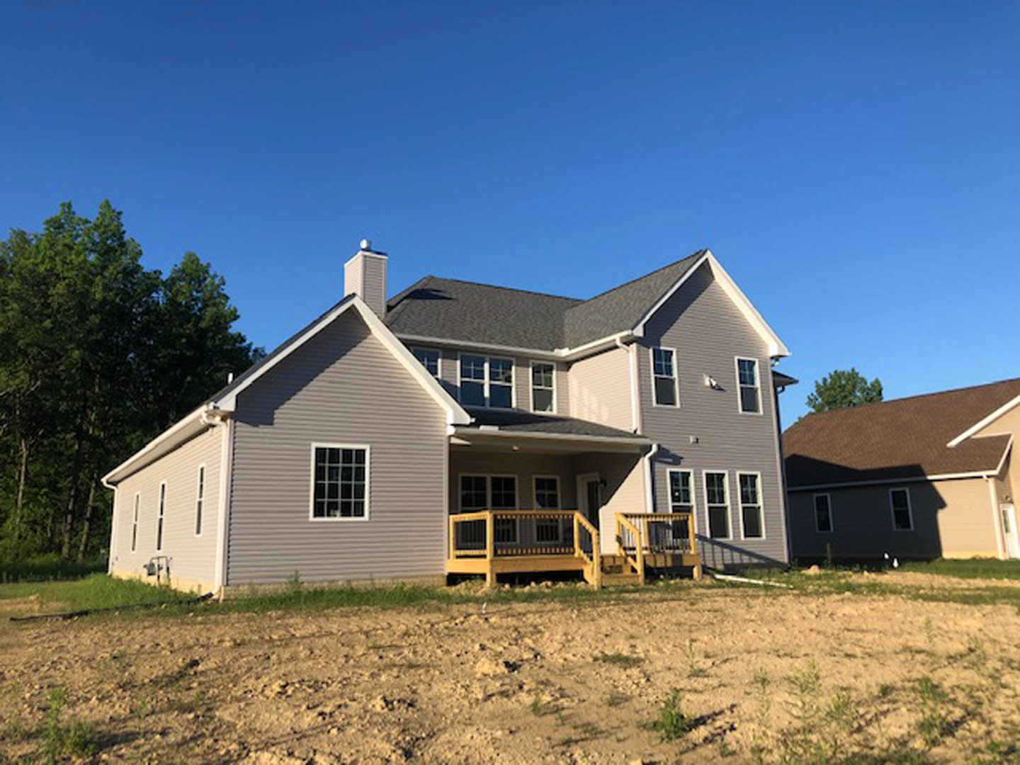 Modern home with brown roof, large grid windows, wooden deck and porch, surrounded by trees, unfinished dirt yard in foreground