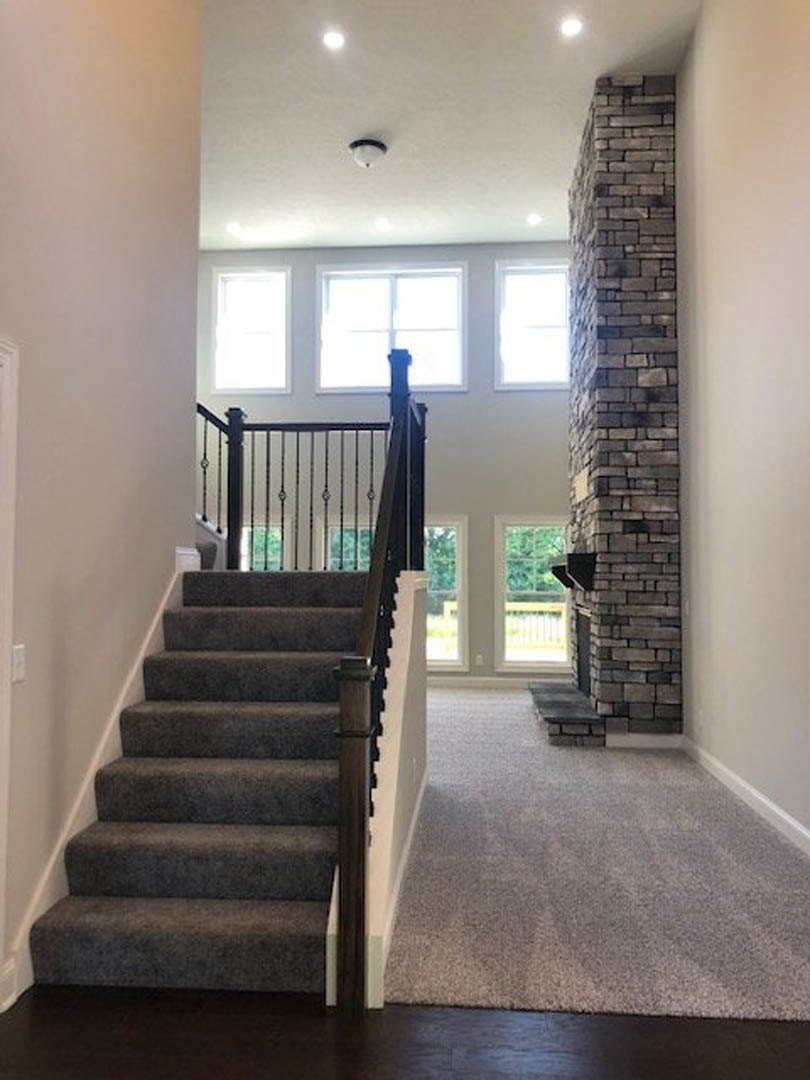 Staircase with black metal railing beside exposed brick wall, carpeted living area with fireplace and large window letting in natural light
