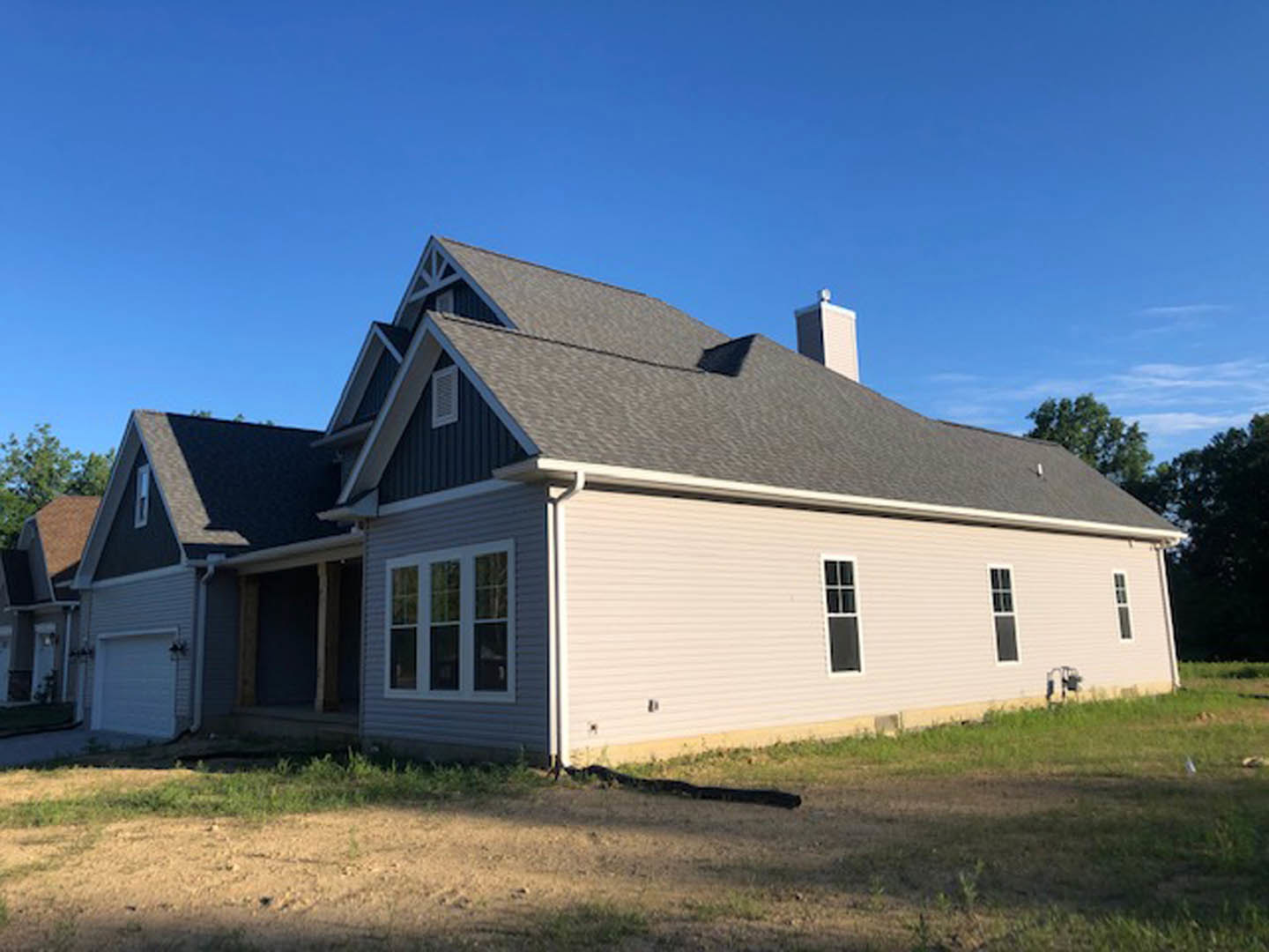Two-story house with gray siding, white trim, brick chimney, large windows, attached white garage door, green lawn, mature trees, and blue sky