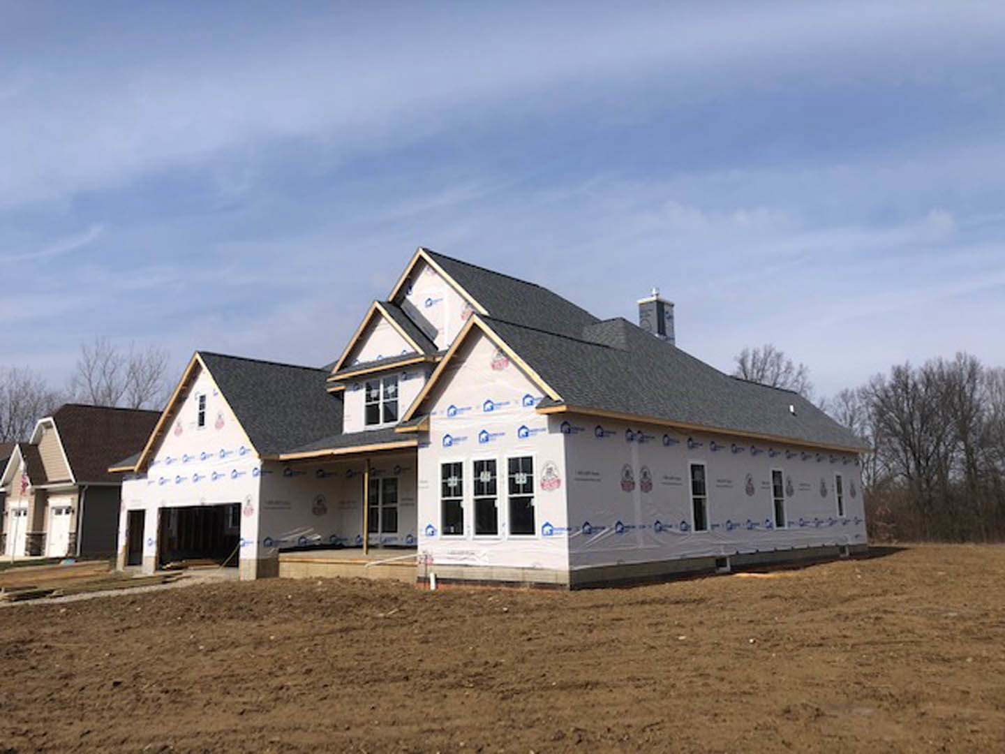 Wood-framed house under construction on a dirt lot, blue sky overhead, distant trees, unfinished windows, American flag mounted near entry