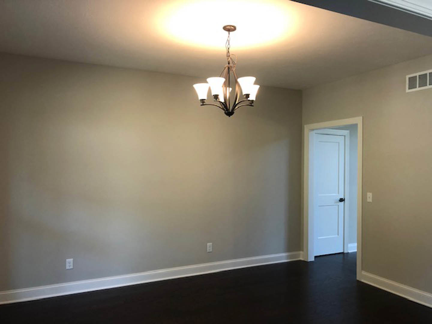 Chandelier with glass light fixtures hangs from a white plaster ceiling above black carpet flooring; white door with black handle and window with white frame visible in room.