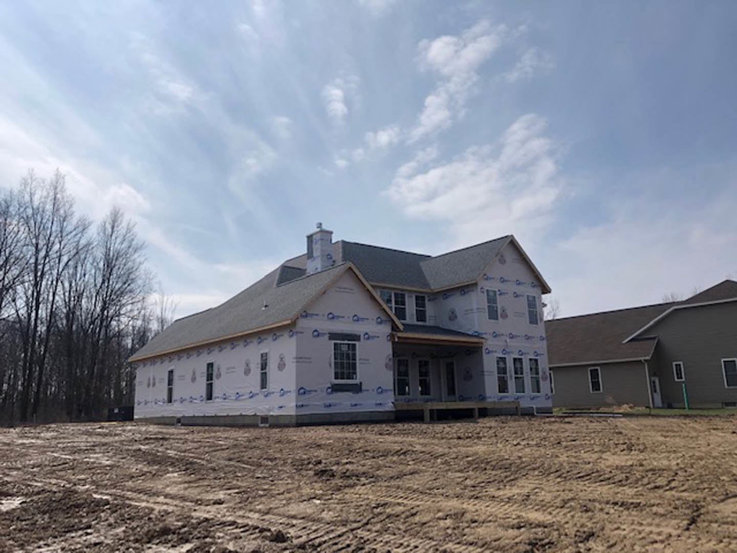 Grey house under construction with exposed insulation, white door, square-paned window, dirt field with tire tracks, and trees in the background under cloudy sky