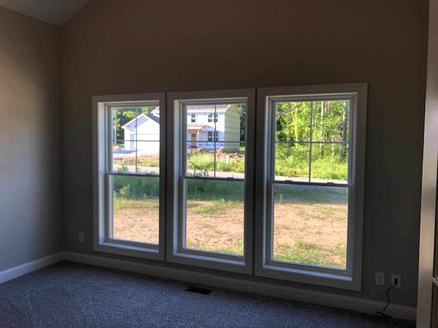 Bright room with three large windows, white walls, hardwood floor, views of green lawn and trees outside, window blinds partially open