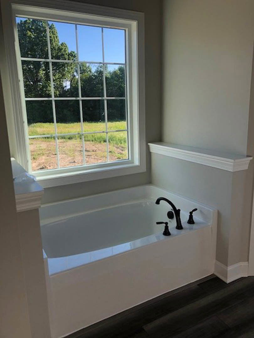Freestanding white bathtub beneath large window, chrome faucet, white walls with molding, view of green trees and field outside