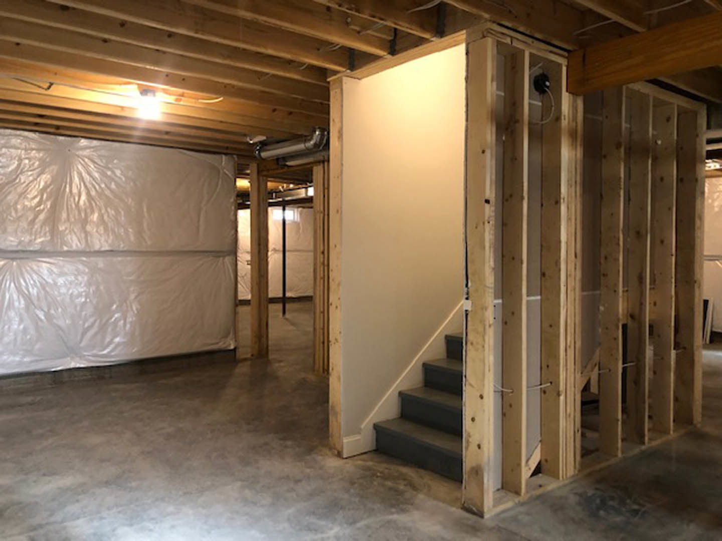 Basement with white plaster walls, exposed wooden ceiling beams with recessed lights, unfinished wooden staircase, and white plastic bag hanging on wall