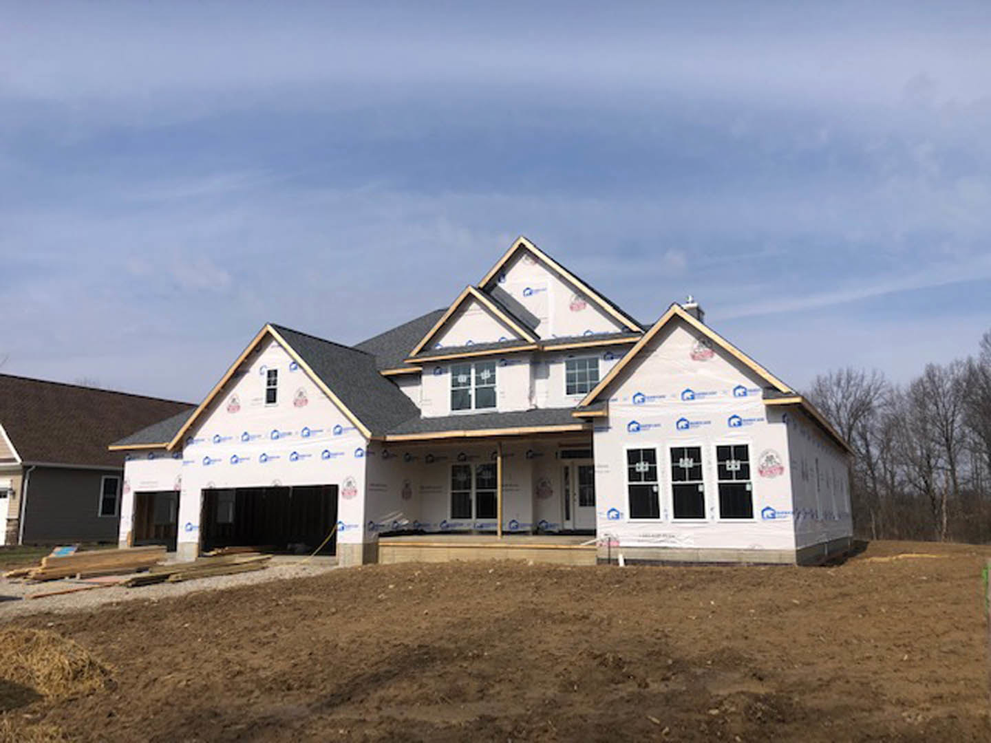 Wood-framed house under construction with multi-pane windows, unfinished roof, dirt field in foreground, and trees in background under blue sky