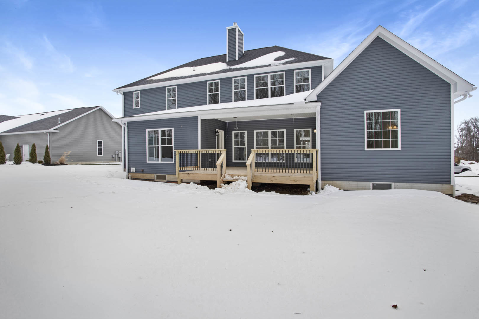 Two-story home with white-framed windows, snow covering the roof and ground, wooden deck and porch with metal railings, winter sky in the background