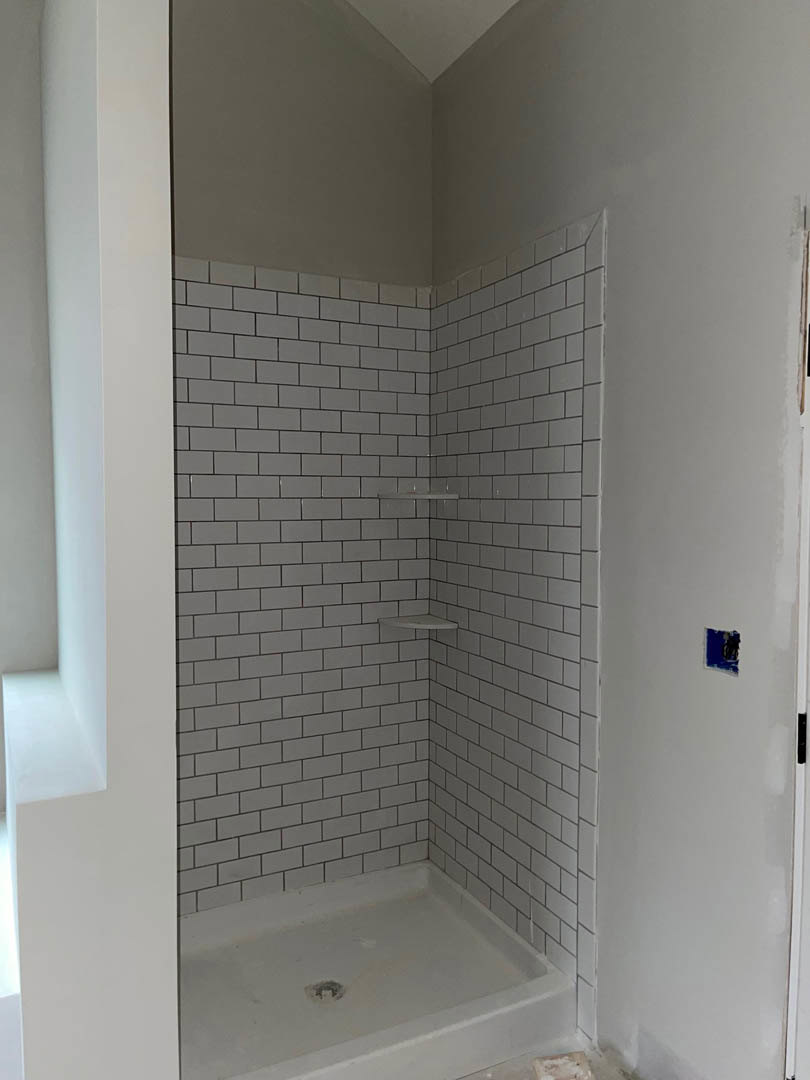 White tile shower wall with built-in bathtub, blue electrical outlet on adjacent wall, black and white object resting on white surface nearby.