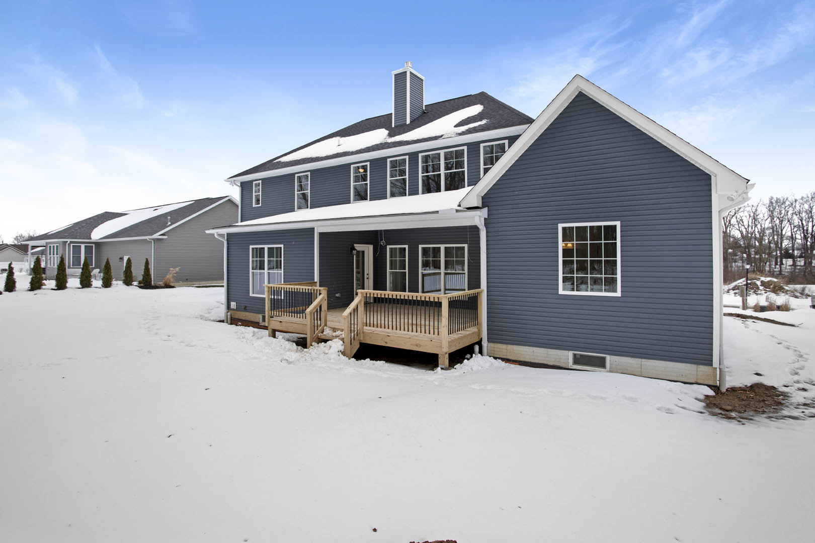 Two-story home with white siding, multi-pane windows, wooden deck featuring black railing, snow covering deck and ground, overcast winter sky in background