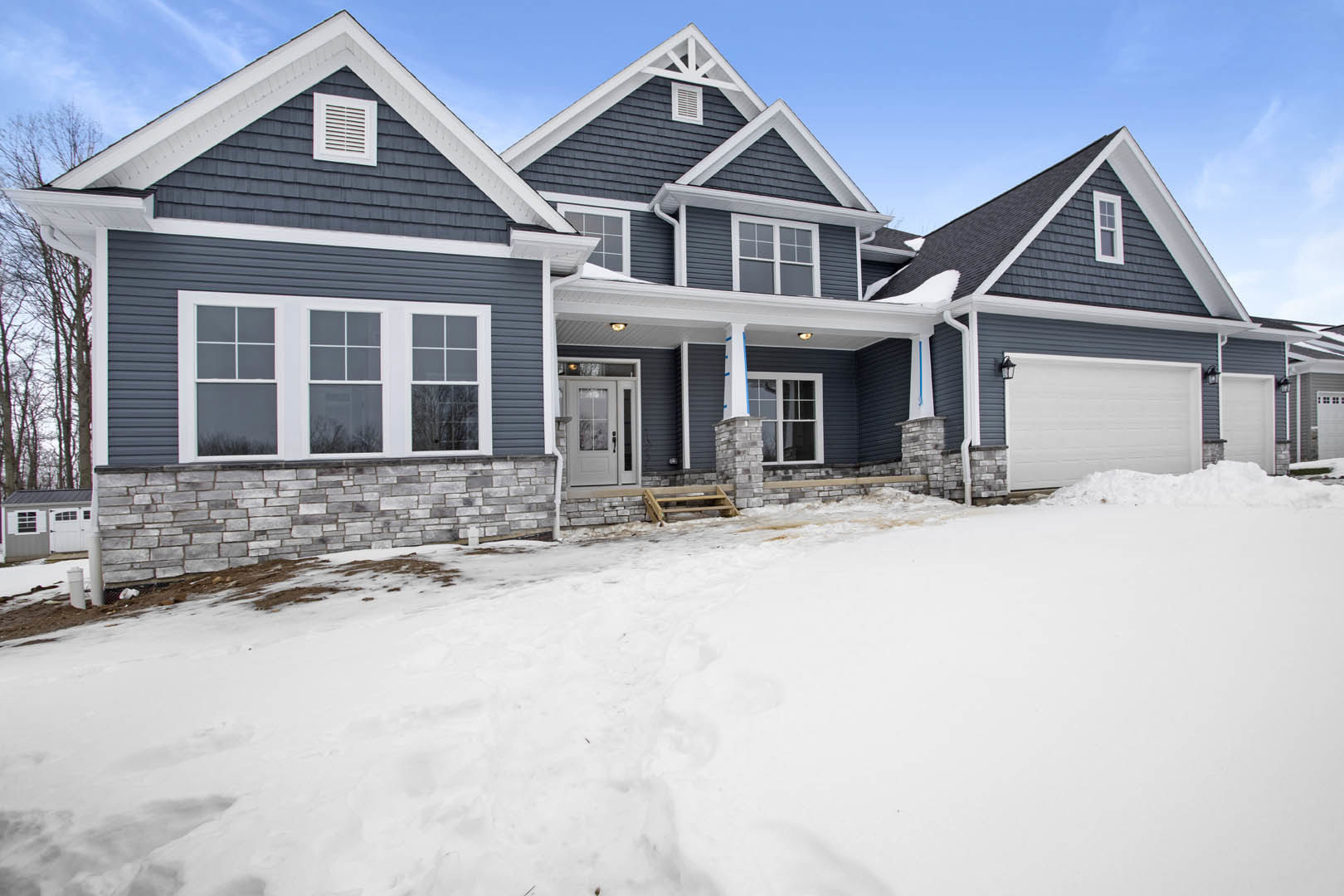 White house with blue roof, snow covering the ground, glass-paneled white front door, garage door partially surrounded by snow, winter landscape with clear sky