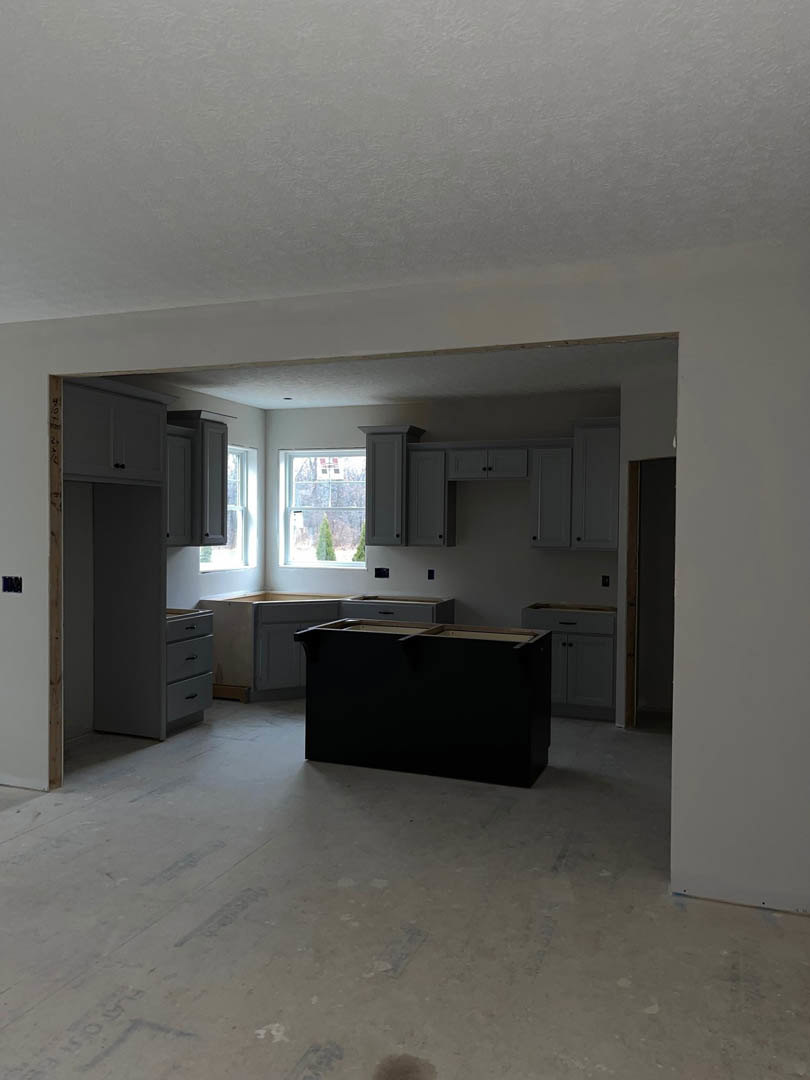 Black kitchen island with matte cabinetry, open wooden shelves, large window overlooking trees, light plaster walls, wood flooring, and recessed ceiling lighting.